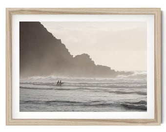 May include: A framed wall art print featuring a monochromatic landscape of a misty coastline with large dark cliffs and ocean waves. Two small figures are visible in the water with a surfboard, set within a light-toned wooden frame.