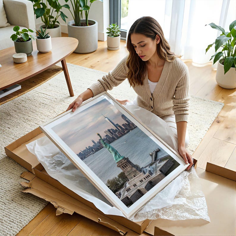 May include: A rectangular framed wall art print featuring an aerial view of the Statue of Liberty and the New York City skyline. The print is encased in a light-colored wooden frame with a white mat border, showcasing the green statue and city.