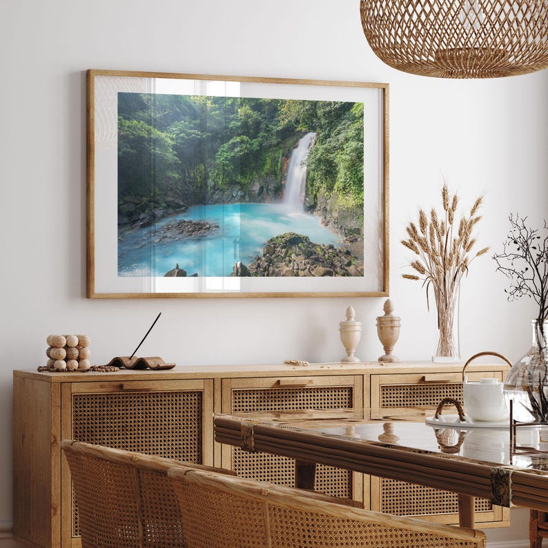 a serene indoor setting with a large framed picture of a waterfall hanging on the wall above a wooden cabinet. The cabinet has a glass top and is situated in a dining area with a table and chairs. The overall scene conveys a sense of tranquility and natural beauty.