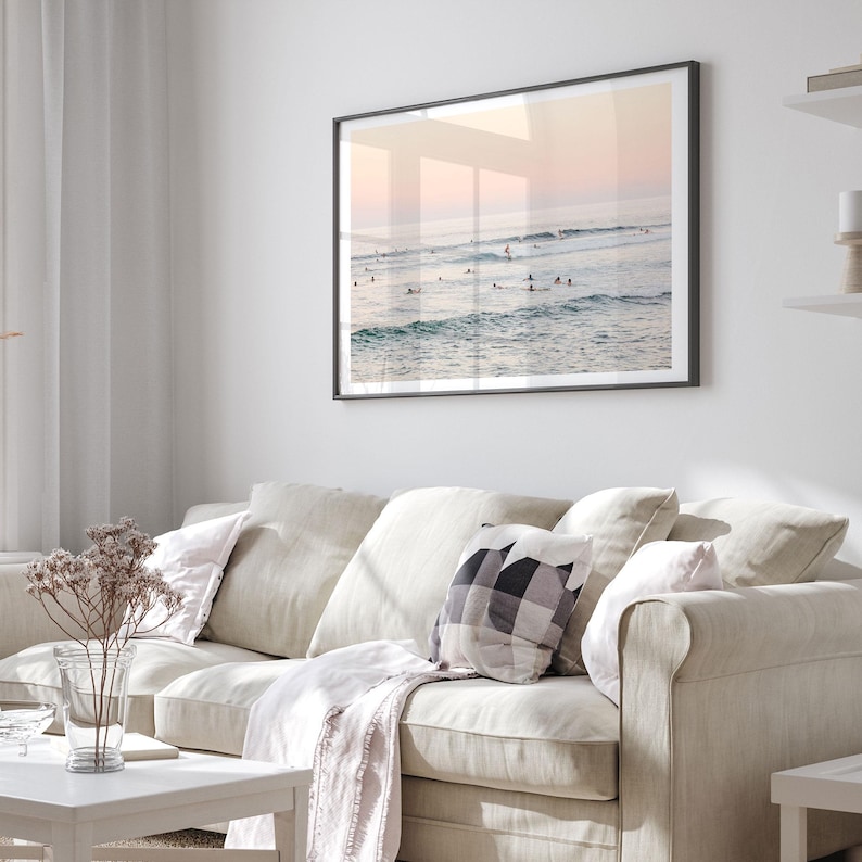 a cozy living room with a beige sofa adorned with a checkered throw pillow and a white blanket draped over it. Above the sofa, a framed picture of people surfing on a beach is displayed on the wall, creating a serene and inviting atmosphere.