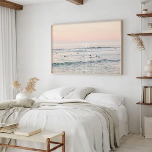 May include: A large rectangular wall art print depicting a calm ocean scene with surfers at sunset, featuring soft pink and blue hues. The print is displayed above a white bed with layered bedding and a wooden bench in a bright room.