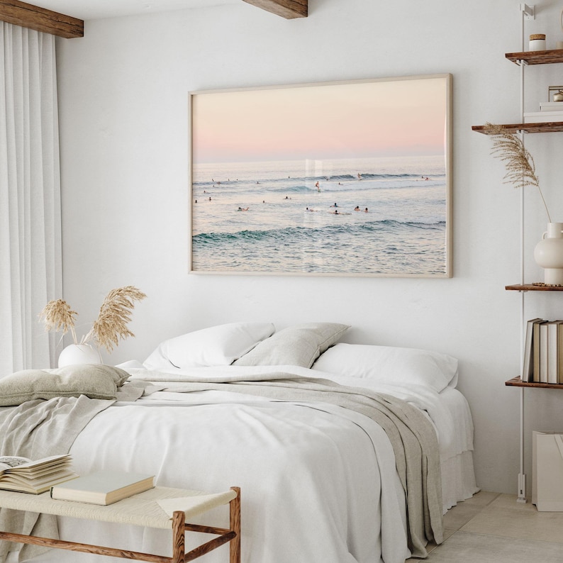 May include: A large rectangular wall art print depicting a calm ocean scene with surfers at sunset, featuring soft pink and blue hues. The print is displayed above a white bed with layered bedding and a wooden bench in a bright room.