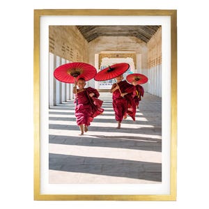 May include: A framed photograph featuring three young individuals in maroon robes running through a stone corridor. Each person holds a vibrant red paper umbrella above their head. The scene is captured in bright daylight, casting long shadows on the floor.
