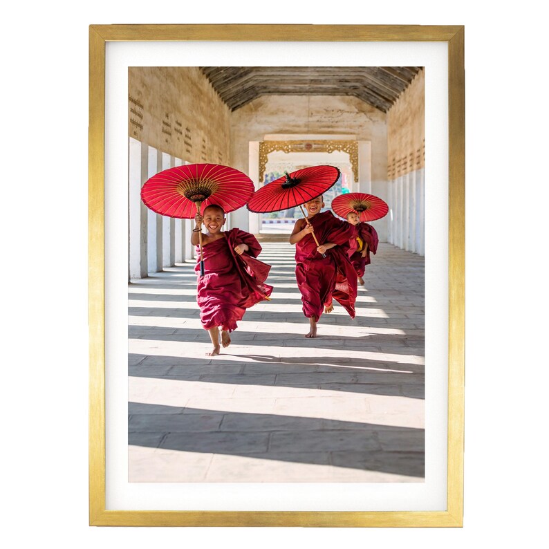 May include: A framed photograph featuring three young individuals in maroon robes running through a stone corridor. Each person holds a vibrant red paper umbrella above their head. The scene is captured in bright daylight, casting long shadows on the floor.