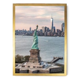 May include: A framed wall art print featuring the Statue of Liberty standing on its pedestal in New York Harbor, with the Manhattan skyline and One World Trade Center visible in the background under a soft, pale sky at dusk or dawn.