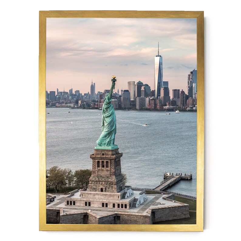 May include: A framed wall art print featuring the Statue of Liberty standing on its pedestal in New York Harbor, with the Manhattan skyline and One World Trade Center visible in the background under a soft, pale sky at dusk or dawn.