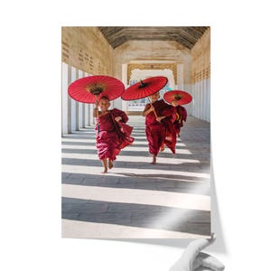 May include: Three young individuals dressed in traditional maroon robes running along a stone corridor, each holding a large, vibrant red paper umbrella. The scene captures a sense of movement and joy as they navigate the sunlit, arched walkway.