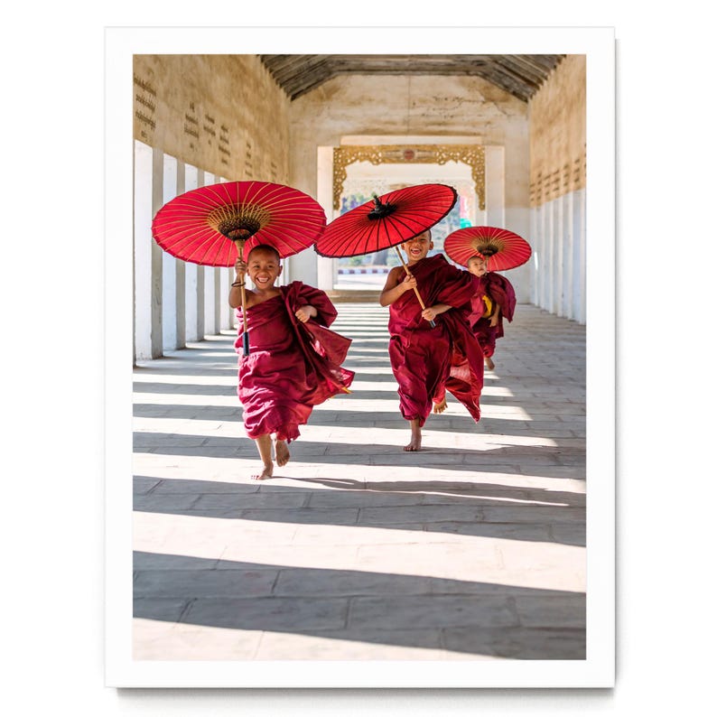 May include: Three young individuals dressed in traditional maroon robes are running along a stone corridor. Each person holds a large, vibrant red paper umbrella overhead. The scene is captured in a bright, sunlit hallway with stone pillars.