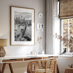 a modern and minimalist living space with a large framed black and white photograph of the Brooklyn Bridge hanging on the wall above a white desk. The desk has a wicker chair and a small plant on it. There is also a clock and a bird figurine on the desk.