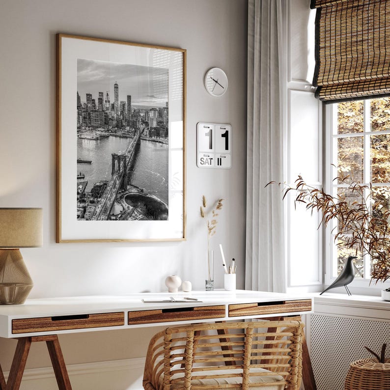 a modern and minimalist living space with a large framed black and white photograph of the Brooklyn Bridge hanging on the wall above a white desk. The desk has a wicker chair and a small plant on it. There is also a clock and a bird figurine on the desk.