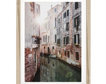May include: A framed wall art print featuring a narrow canal in Venice, Italy, with weathered brick buildings on both sides, a small boat floating on the water, and bright sunlight creating a starburst effect above the historic architecture.