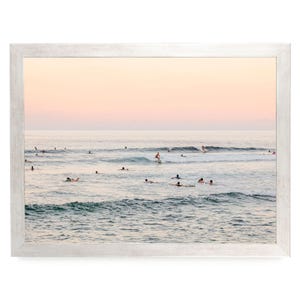 May include: A framed landscape photograph of an ocean scene at sunset with a soft pink and orange sky. Numerous surfers are scattered across the water, with one individual actively riding a wave on a surfboard amidst the gentle rolling swells.