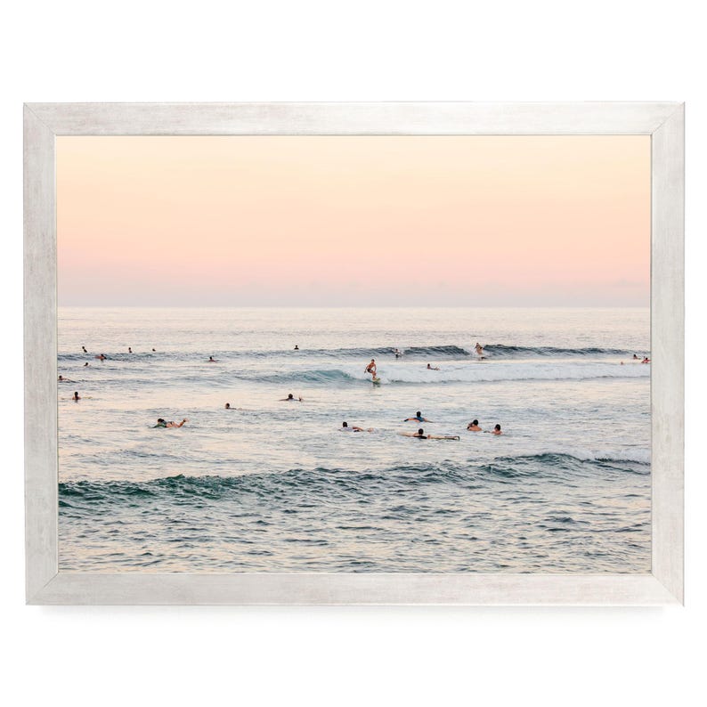 May include: A framed landscape photograph of an ocean scene at sunset with a soft pink and orange sky. Numerous surfers are scattered across the water, with one individual actively riding a wave on a surfboard amidst the gentle rolling swells.