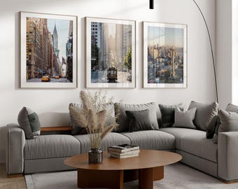 May include: Three framed wall art prints featuring cityscapes of New York, San Francisco, and Seattle, displayed above a gray sectional sofa with a wooden oval coffee table holding a vase of dried pampas grass and a stack of books.