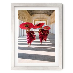 May include: A framed photograph showing three young individuals in maroon robes running along a stone corridor. Each person holds a vibrant red paper umbrella overhead. The scene is captured in bright daylight, casting long shadows on the ground.