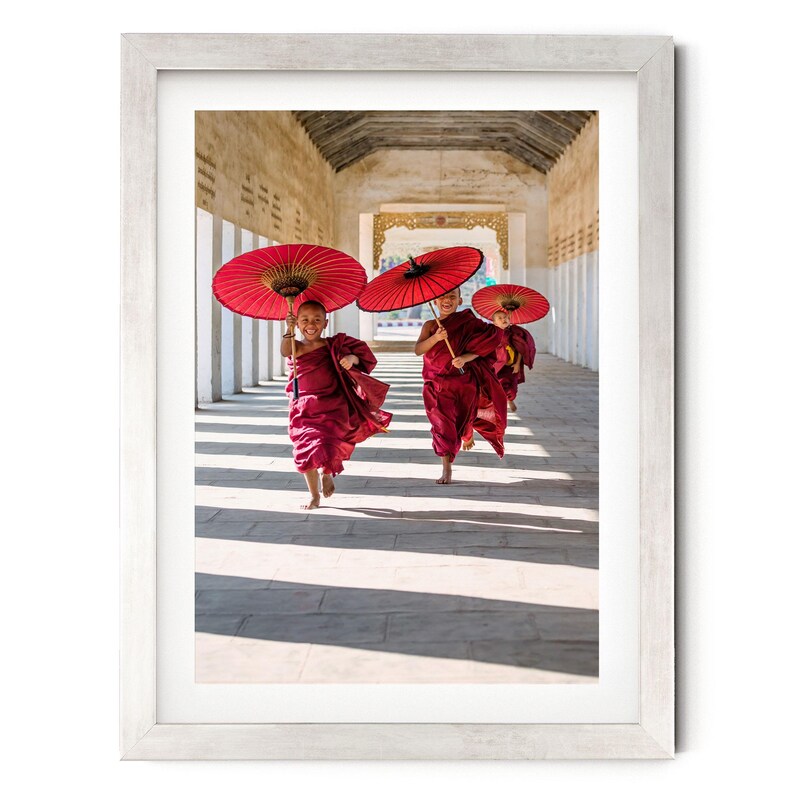 May include: A framed photograph showing three young individuals in maroon robes running along a stone corridor. Each person holds a vibrant red paper umbrella overhead. The scene is captured in bright daylight, casting long shadows on the ground.