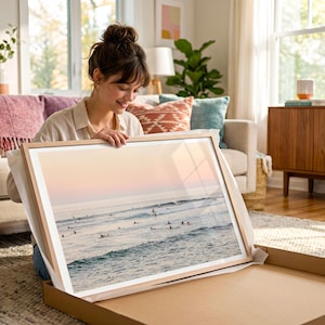 A woman is sitting on the floor, holding a framed picture of a beach scene, which appears to be a collage of multiple images.