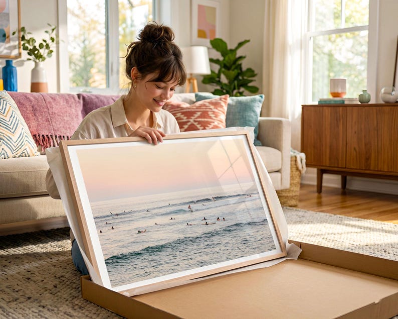 A woman is sitting on the floor, holding a framed picture of a beach scene, which appears to be a collage of multiple images.