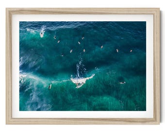 May include: A framed aerial photograph of a deep blue ocean with multiple surfers on surfboards scattered across the water. One surfer is riding a breaking wave, creating white foam, while others wait in the surrounding dark turquoise sea.