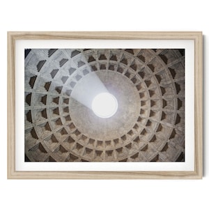 May include: A framed print featuring the interior of a domed ceiling with a central oculus. The ceiling is constructed of gray stone blocks arranged in a concentric pattern. A beam of light shines through the oculus, illuminating the center.