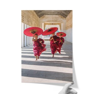 May include: Three young individuals wearing deep red robes are running along a stone corridor. Each person holds a large, vibrant red paper umbrella with a wooden handle. The scene is captured in a bright, sunlit hallway with stone pillars.