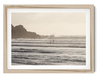May include: A framed wall art print featuring a sepia-toned coastal landscape with ocean waves, a rocky shoreline, and surfers in the water. The frame is made of light-colored wood with a visible grain, surrounding a white mat and the photograph.