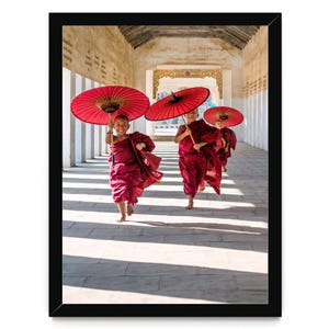 May include: A framed photograph showing three individuals in maroon robes running along a stone corridor. Each individual holds a vibrant red paper umbrella overhead. The scene is captured in a long, light-filled hallway with white pillars.