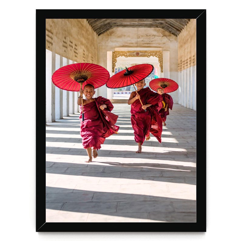 May include: A framed photograph showing three individuals in maroon robes running along a stone corridor. Each individual holds a vibrant red paper umbrella overhead. The scene is captured in a long, light-filled hallway with white pillars.