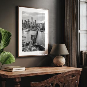 a framed black and white photograph of the Brooklyn Bridge hanging on a wall above a wooden desk with a lamp and a book.