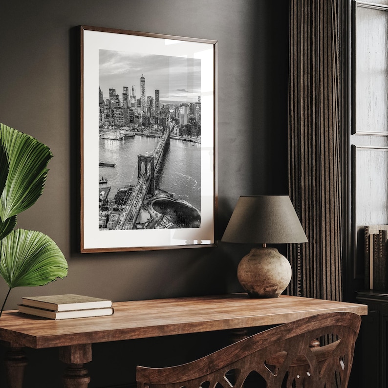 a framed black and white photograph of the Brooklyn Bridge hanging on a wall above a wooden desk with a lamp and a book.