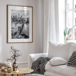 a living room with a large framed black and white photograph of the Brooklyn Bridge hanging on the wall. The photograph captures the bridge and its surroundings, creating a focal point in the room. The living room is furnished with a white sofa, a wooden coffee table, and a vase with branches. The overall decor and arrangement of the room create a cozy and inviting atmosphere.