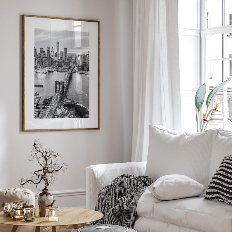 a living room with a large framed black and white photograph of the Brooklyn Bridge hanging on the wall. The photograph captures the bridge and its surroundings, creating a focal point in the room. The living room is furnished with a white sofa, a wooden coffee table, and a vase with branches. The overall decor and arrangement of the room create a cozy and inviting atmosphere.
