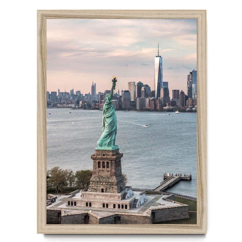 May include: A framed wall art print featuring the Statue of Liberty in New York Harbor, shown from behind with the Manhattan skyline in the background. The statue is verdigris green, set against a light wood-toned frame under a soft sky.