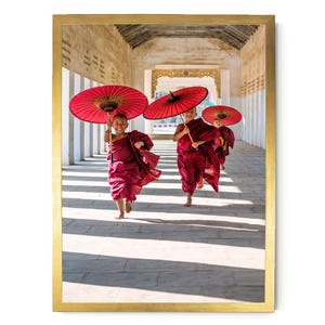 May include: A framed photograph shows three young individuals in maroon robes running through a stone corridor. Each person holds a vibrant red paper umbrella overhead. The scene is captured in a long, sunlit hallway with white pillars.