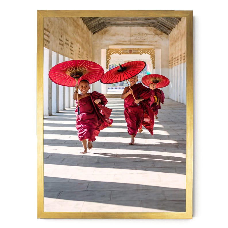 May include: A framed photograph shows three young individuals in maroon robes running through a stone corridor. Each person holds a vibrant red paper umbrella overhead. The scene is captured in a long, sunlit hallway with white pillars.