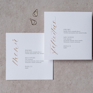 Könnte beinhalten: Zwei quadratische weiße Menükarten mit goldener Kalligraphie und einer Liste von Gerichten. Auf einer Karte steht das Wort "Menü", auf der anderen "Felicitas". Goldene Büroklammern sind im Hintergrund. Das Menü umfasst "Primi", "Secondi" und "Dolci".