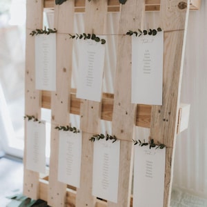 Könnte beinhalten: Eine Holzpalette, dekoriert mit Grün und weißen Karten. Die Karten sind mit Schnur befestigt und zeigen Namen, wahrscheinlich für eine Hochzeit oder Veranstaltung. Eukalyptusblätter sind oben und unten angeordnet und verleihen dem rustikalen Display eine natürliche Note.