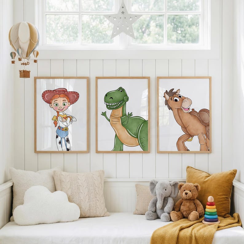 May include: Three framed art prints featuring cartoon characters: a cowgirl, a green dinosaur, and a brown horse. The prints are displayed above a white daybed with decorative pillows and stuffed animals. The room has a window and a star-shaped decoration.