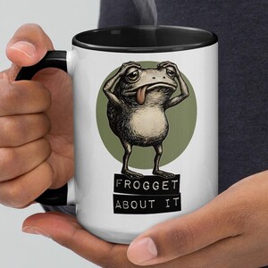 Op de afbeelding: Een witte 425 ml keramische mok met een zwarte binnenkant en handvat. De mok heeft een verweerde illustratie van een kikker met zijn tong uit, tegen een groene cirkel. De tekst "FROGGET ABOUT IT" staat eronder.