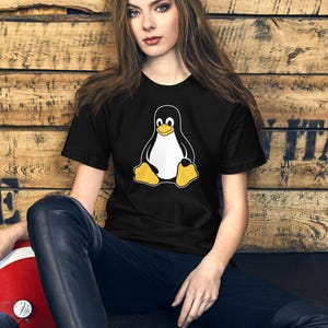 May include: Black t-shirt featuring the Linux penguin mascot in white, black, and yellow. The person is wearing black pants and is sitting in front of a wooden background. A red and white helmet is visible.