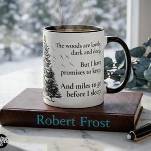 May include: An 11 oz white coffee mug with a black handle and rim, featuring a quote from Robert Frost's poem. The mug has a tree illustration and text: "The woods are lovely, dark and deep..." It sits on a brown book.