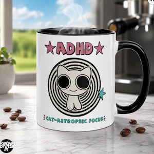 May include: White ceramic coffee mug with a black interior and handle. The mug features the text "ADHD" in pink with star accents, and a cartoon cat illustration inside a spiral. The phrase "CAT-ASTROPHIC FOCUS" is printed below the cat. The mug holds 11 oz.