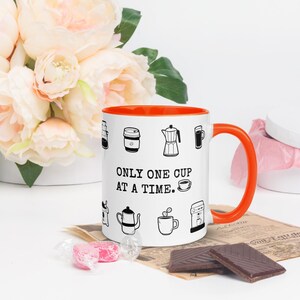 Op de afbeelding: Witte koffiemok met een oranje binnenkant en oor, versierd met zwarte lijntekeningen van koffiezetapparaten. De mok heeft de tekst "ONLY ONE CUP AT A TIME." en is omringd door bloemen en chocolade.