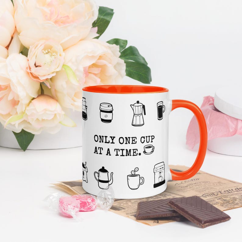 Op de afbeelding: Witte koffiemok met een oranje binnenkant en oor, versierd met zwarte lijntekeningen van koffiezetapparaten. De mok heeft de tekst "ONLY ONE CUP AT A TIME." en is omringd door bloemen en chocolade.