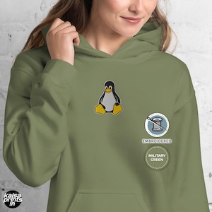 Könnte beinhalten: Militärgrüner Kapuzenpullover mit einem gestickten Pinguin-Motiv auf der Brust. Der Hoodie hat eine Kapuze mit Kordelzug und eine Kängurutasche. Das Design enthält die Wörter "EMBROIDERED" und "MILITARY GREEN".