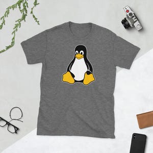 Puede incluir: Camiseta gris jaspeado con un dise&ntilde;o de ping&uuml;ino de dibujos animados. El ping&uuml;ino tiene un cuerpo negro, vientre blanco, pies amarillos y pico amarillo. El ping&uuml;ino est&aacute; sentado. La camiseta es de material suave.