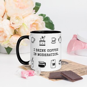 Op de afbeelding: Een witte keramische koffiemok met een zwarte binnenkant en handvat. De mok heeft zwarte lijntekeningen van koffiegerelateerde items en de tekst "I drink coffee in moderation." De mok is omgeven door bloemen en chocolade.