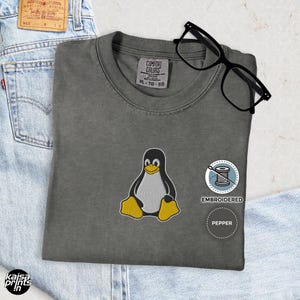 Könnte beinhalten: Graues T-Shirt mit gesticktem Tux-Pinguin-Design. Das Shirt hat einen Rundhalsausschnitt und kurze Ärmel. Das Shirt hat auch ein Etikett mit der Aufschrift "Comfort Colors" und der Größe "XL - TG - EG".