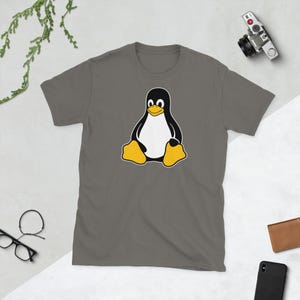Puede incluir: Camiseta gris con un dise&ntilde;o de ping&uuml;ino de dibujos animados. El ping&uuml;ino tiene un cuerpo negro, vientre blanco, pies amarillos y una cara sonriente. La camiseta es de manga corta.
