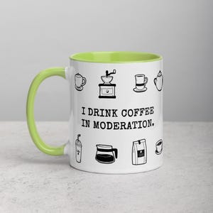 Op de afbeelding: Witte keramische koffiemok met een lichtgroen interieur en handvat. De mok is versierd met zwarte lijntekeningen van koffiegerelateerde items en de tekst "I DRINK COFFEE IN MODERATION."